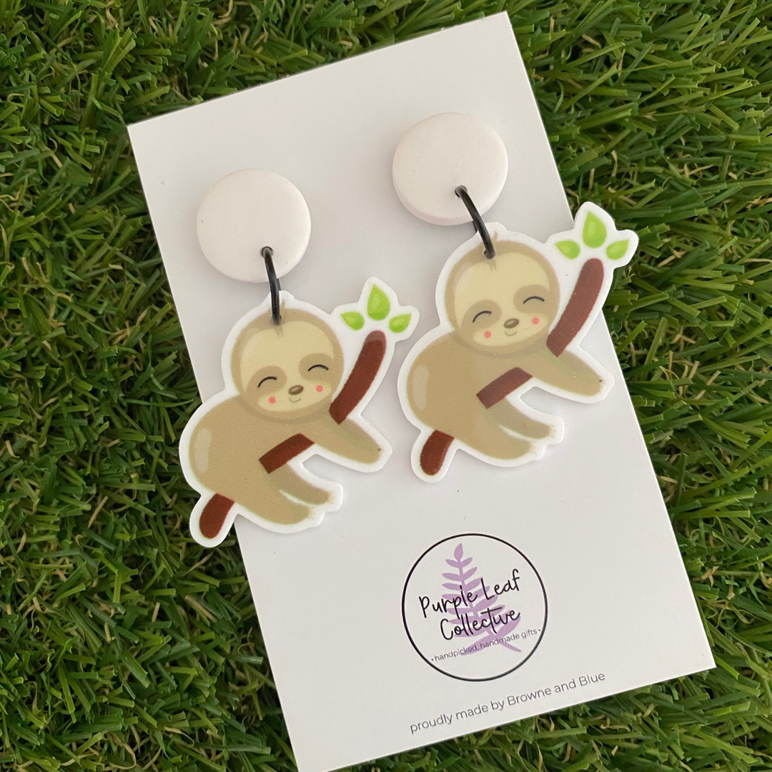 A pair of dangle earrings featuring a pair of sloths resting on tree branches set on crisp white clay tops.