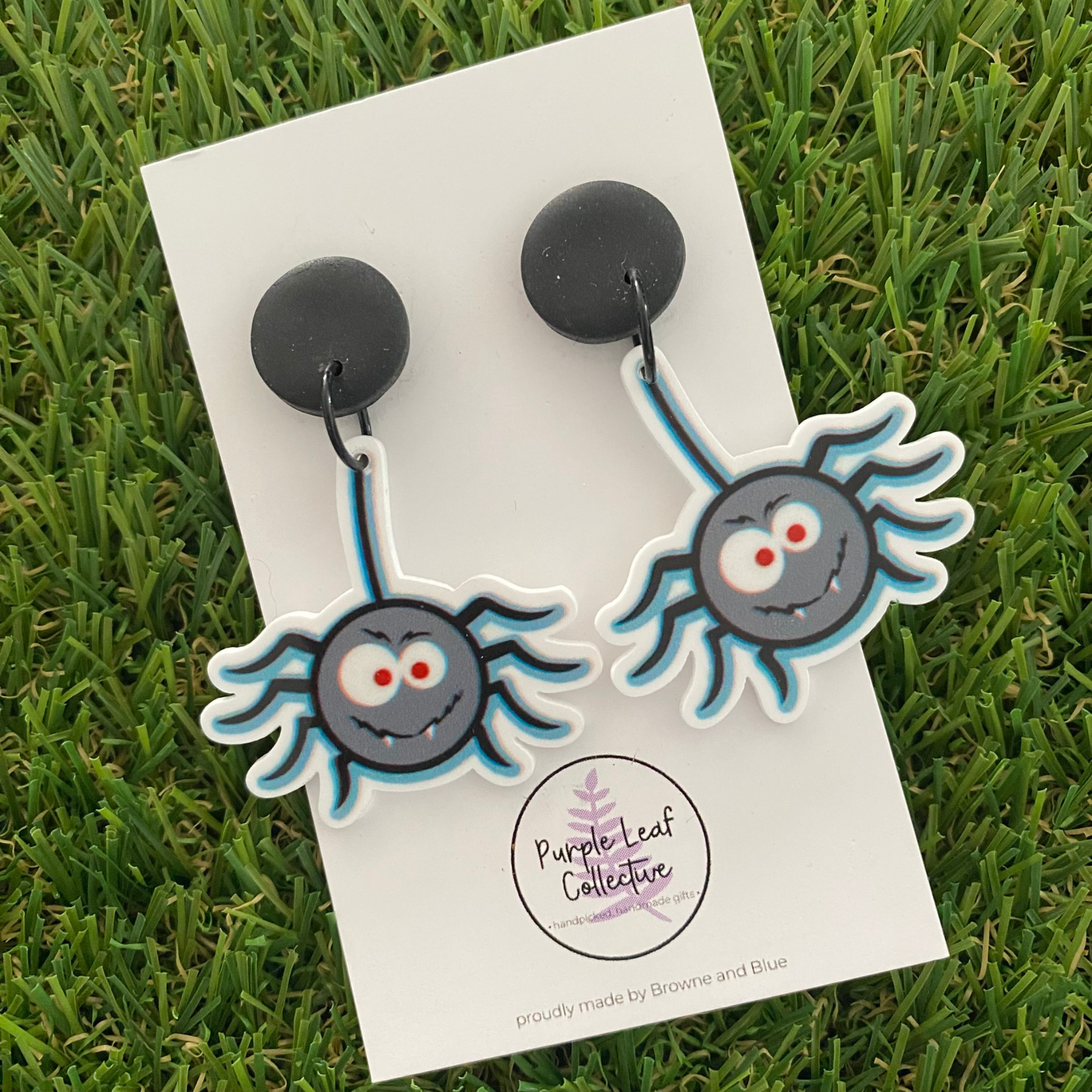 A pair of spider dangle earrings featuring a grey face with blue legs, white outlines and red eyes, set on  black round studs.