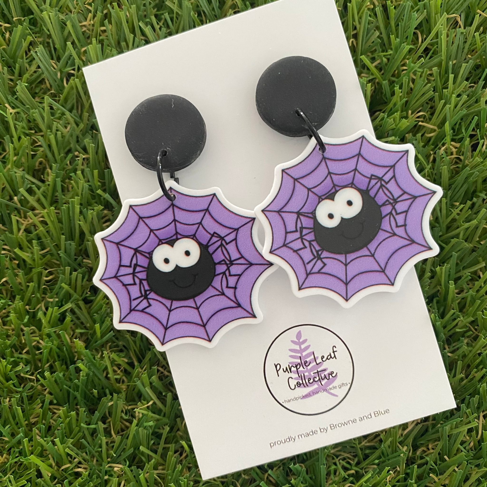 A pair of Halloween spider earrings with purple spiderweb designs, black spider faces sitting on black stud tops.