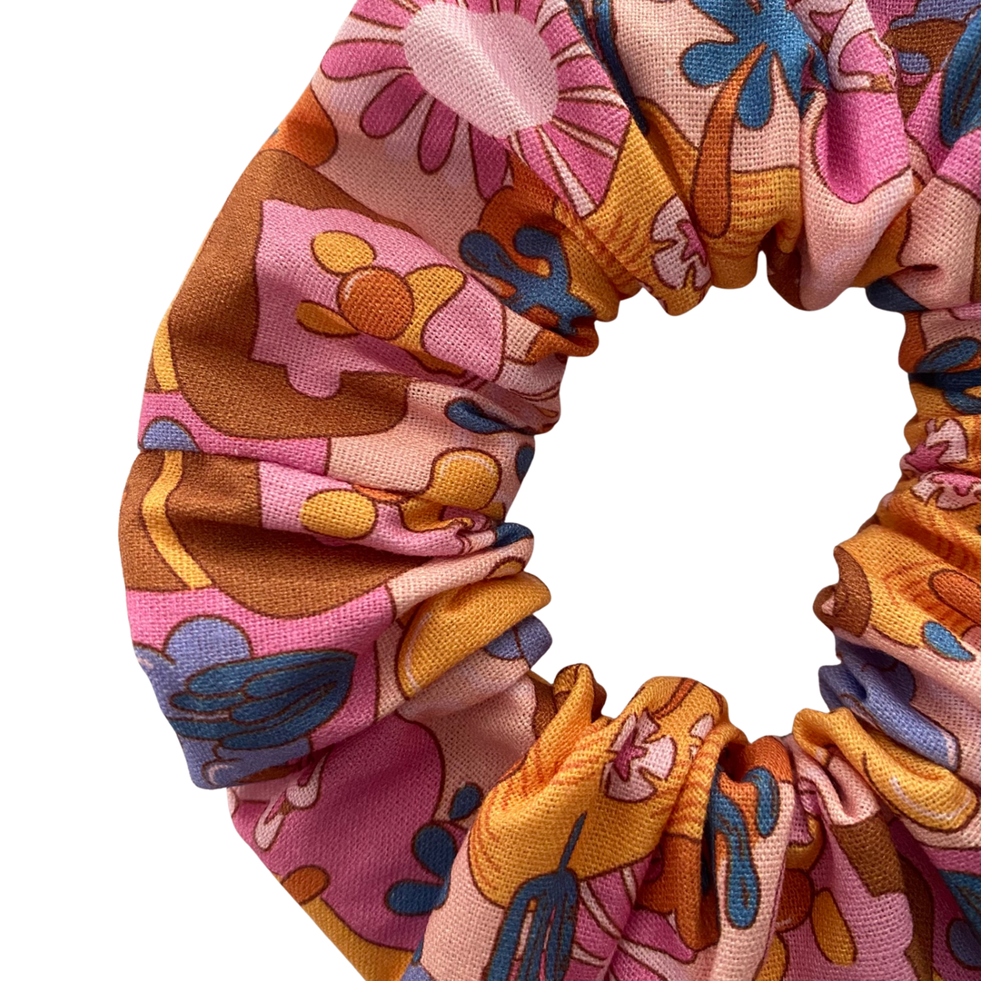 Close up view of a light pink fabric scrunchie featuring a pattern of Bubble O'Bill icons, set against a background of flowers and ranch imagery.