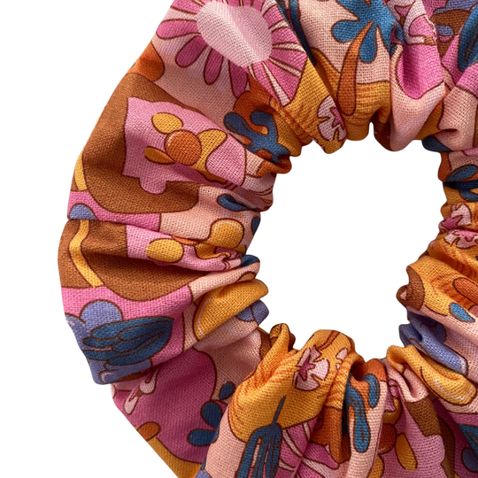 Close up view of a light pink fabric scrunchie featuring a pattern of Bubble O'Bill icons, set against a background of flowers and ranch imagery.