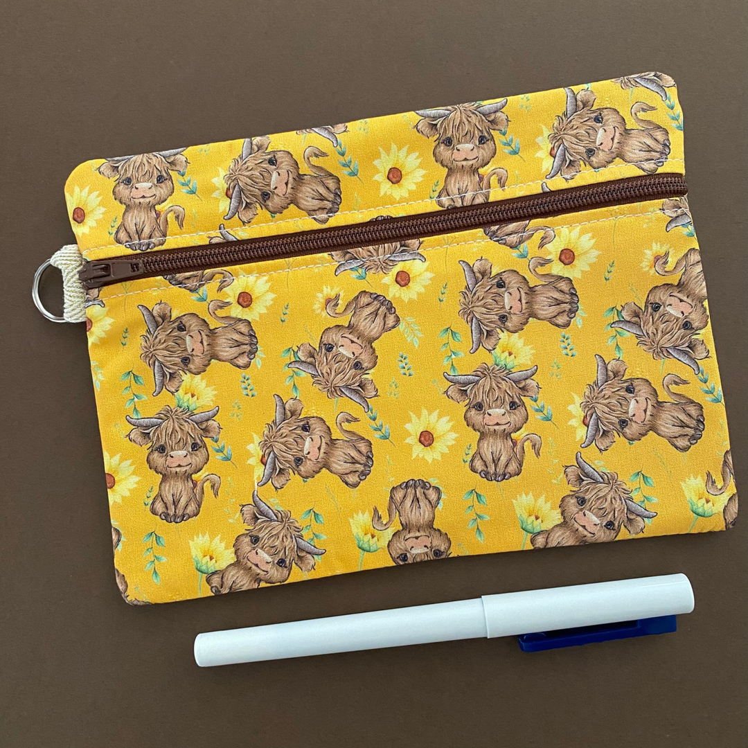 Front view of a zipper pouch with images of baby highland cows and sunflowers on dark yellow fabric.