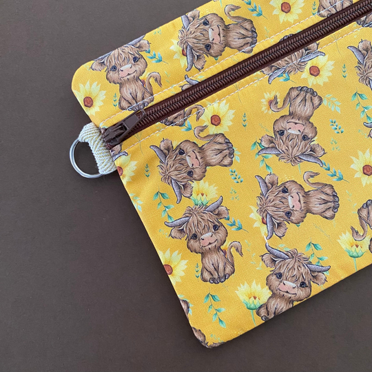 Zipper pouch with images of baby highland cows and sunflowers on dark yellow fabric with clasp.