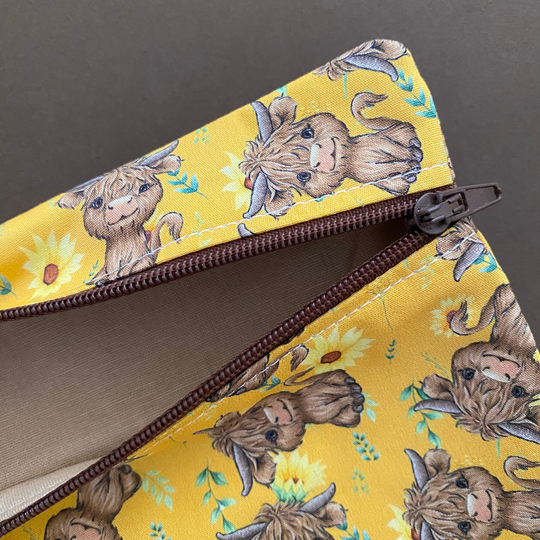 Zipper pouch with images of baby highland cows and sunflowers on dark yellow fabric showing the light brown lining.
