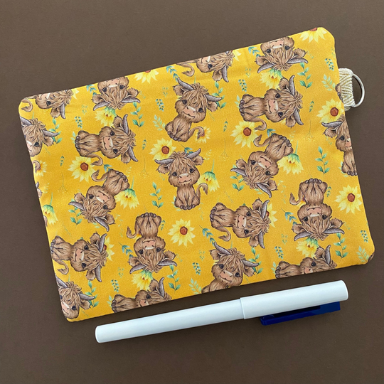 Reverse view of a zipper pouch with images of baby highland cows and sunflowers on dark yellow fabric.