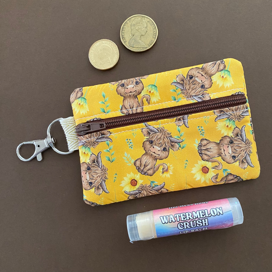 Front view of a mini pouch with images of baby highland cows and sunflowers on dark yellow fabric.