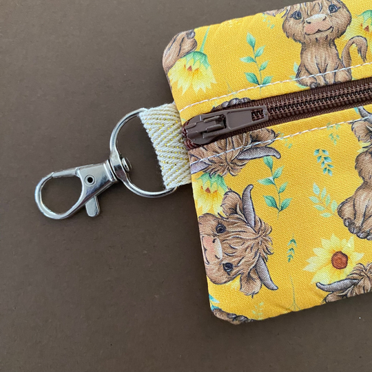 Mini pouch with images of baby highland cows and sunflowers on dark yellow fabric with clip clasp.