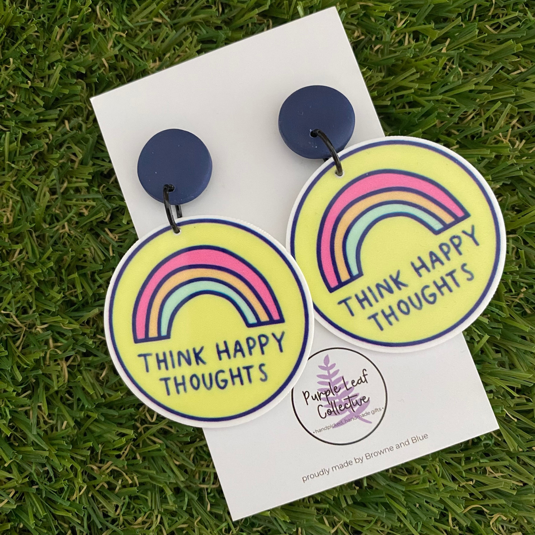 Earrings with rainbow design and 'Think Happy Thoughts' text set on navy blue studs on a white backing card.