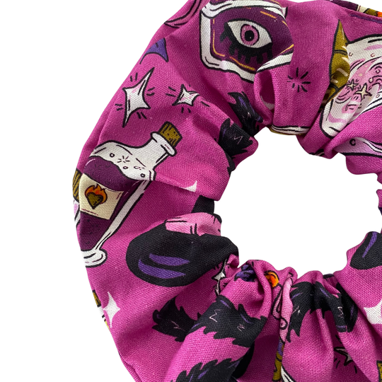Close up view of a purple Halloween scrunchie with a pattern of black cats, bubbling cauldrons, potion bottles and skulls, handmade from woven cotton fabric.