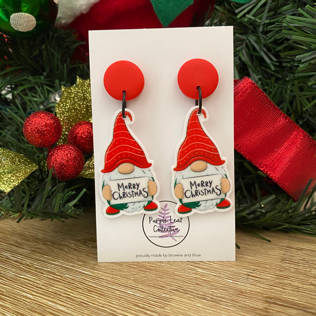 Pair of gnome earrings with 'Merry Christmas' text on a card, surrounded by festive decorations.