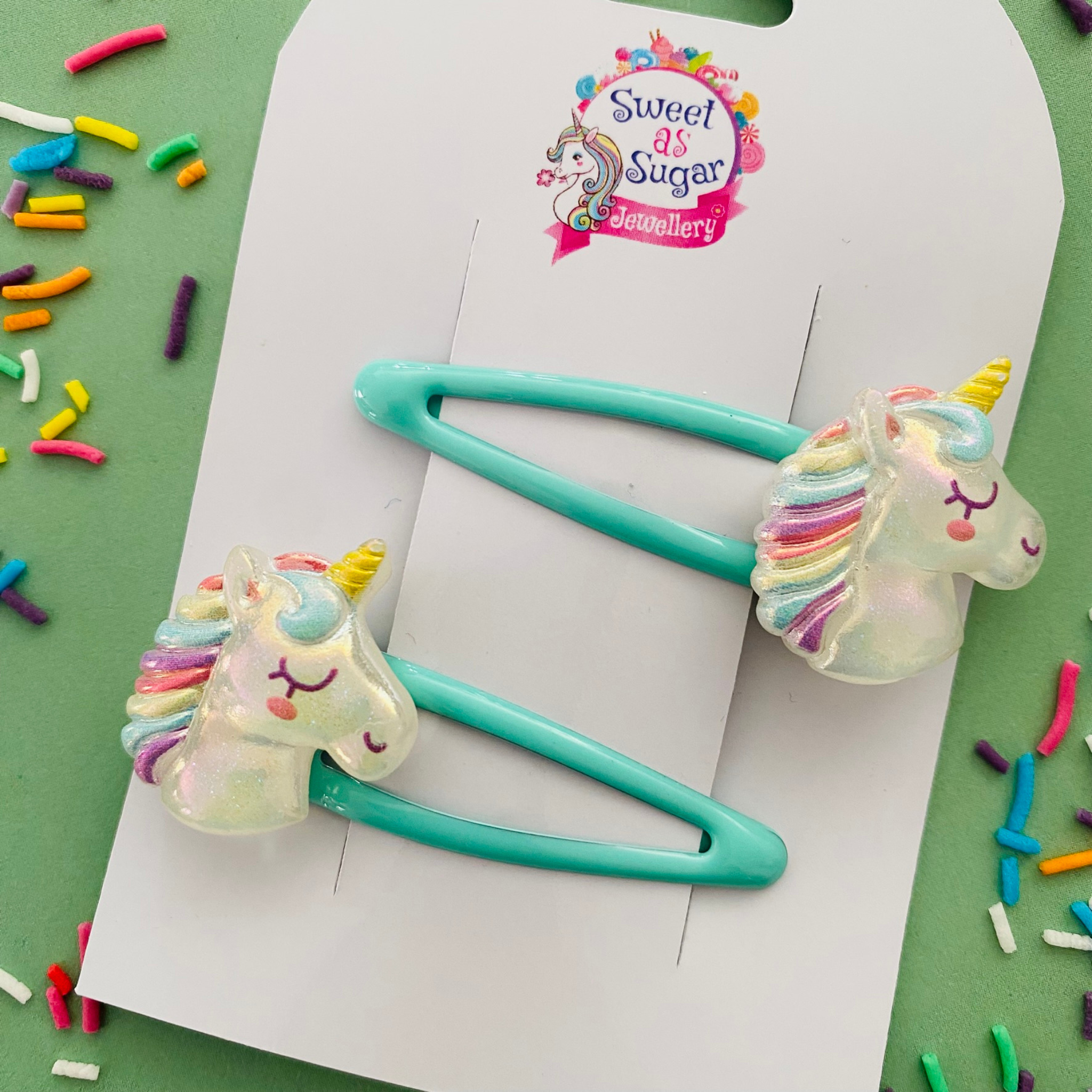 A pair of aqua blue snap clips featuring a unicorn face with a golden horn and rainbow mane.