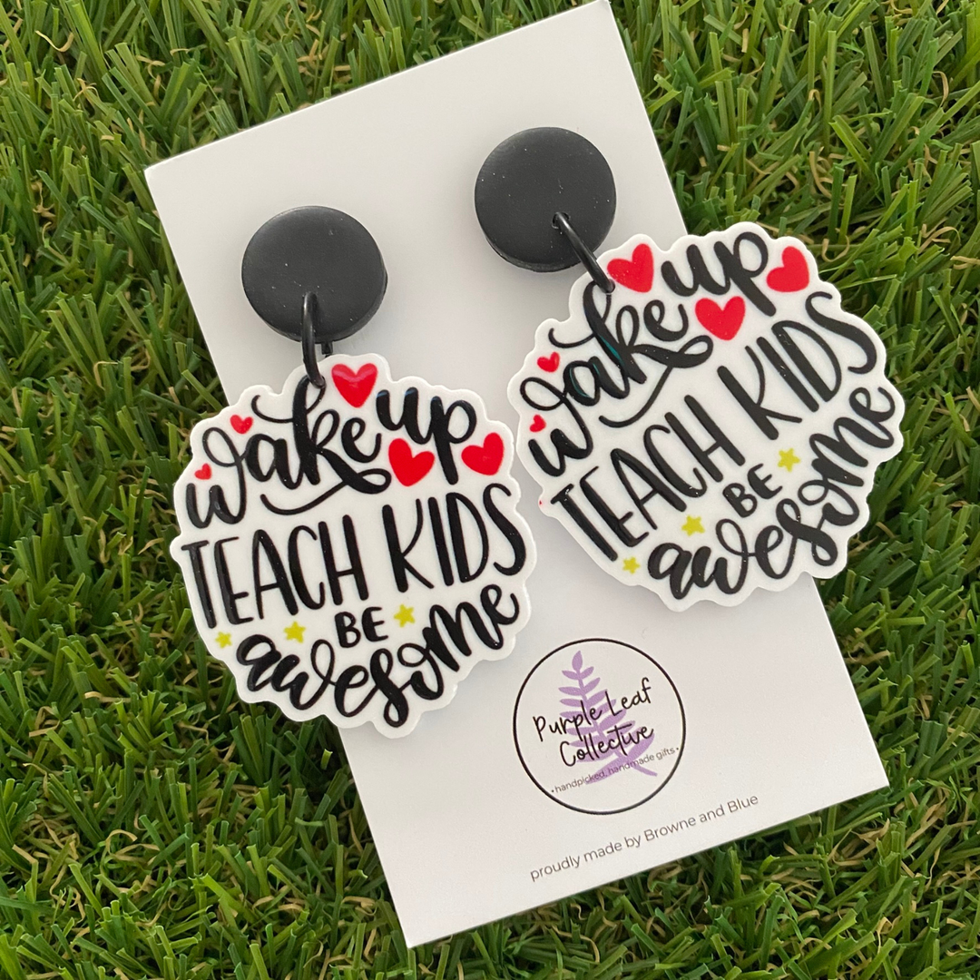 A pair of dangle earrings featuring text 'wake up TEACH KIDS be awesome' in black script with red hearts and small yellow stars, attached to black round studs.