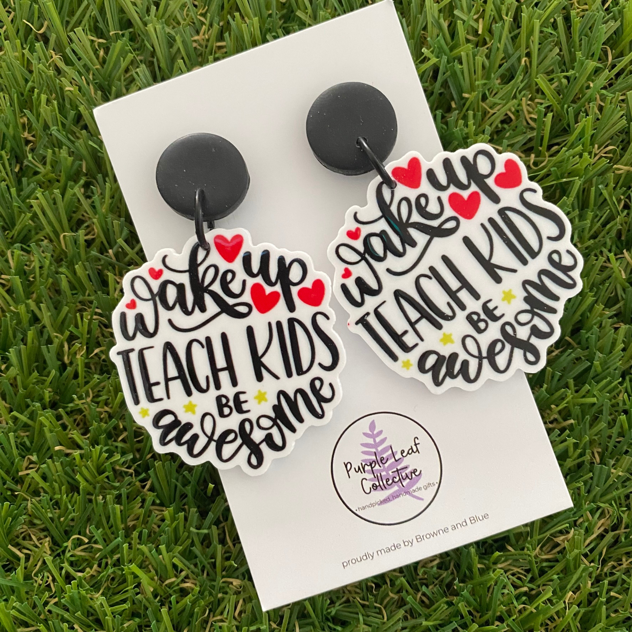 A pair of dangle earrings featuring text 'wake up TEACH KIDS be awesome' in black script with red hearts and small yellow stars, attached to black round studs.
