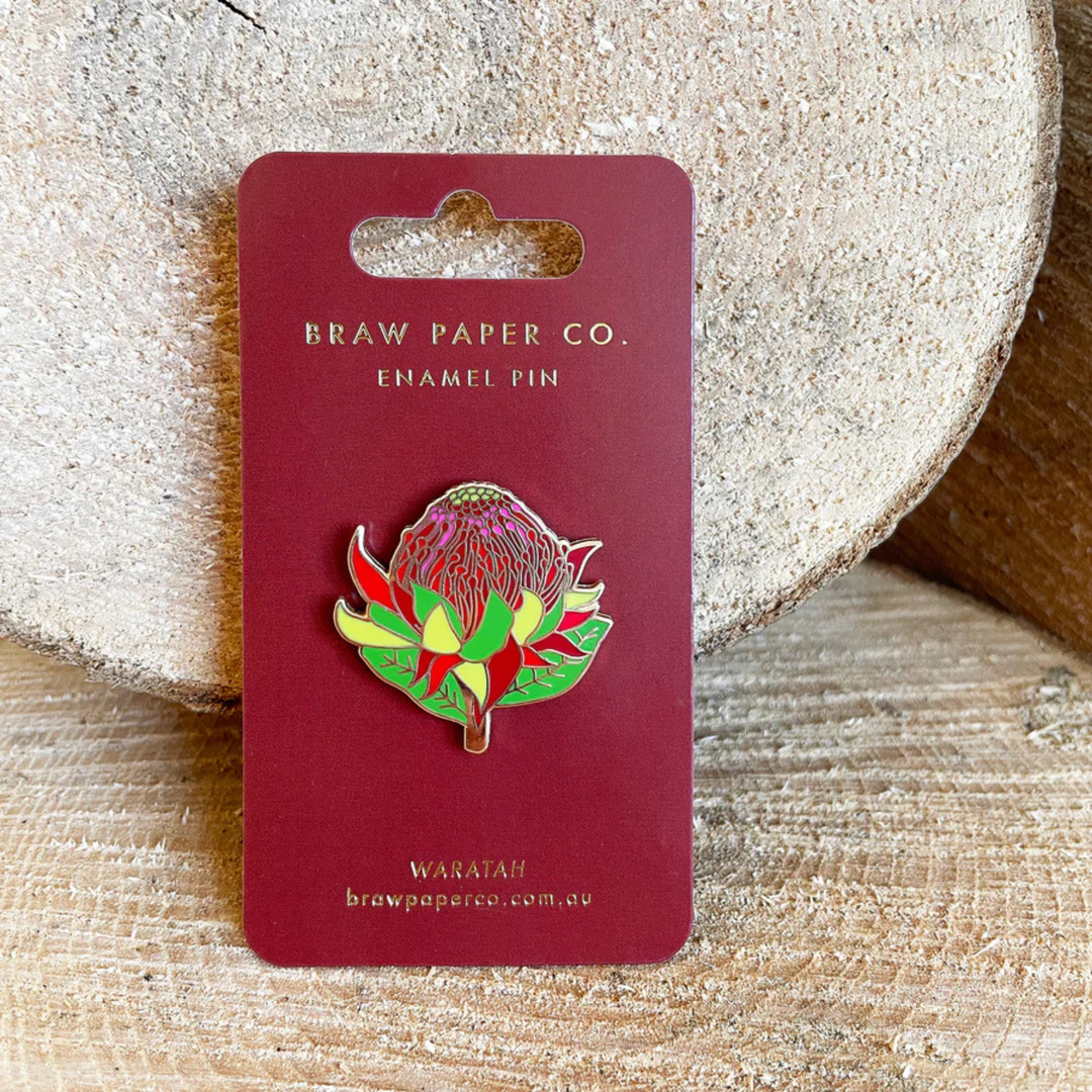 Enamel pin of a waratah flower on a red card with 'Braw Paper Co.' branding, placed on a wooden surface.