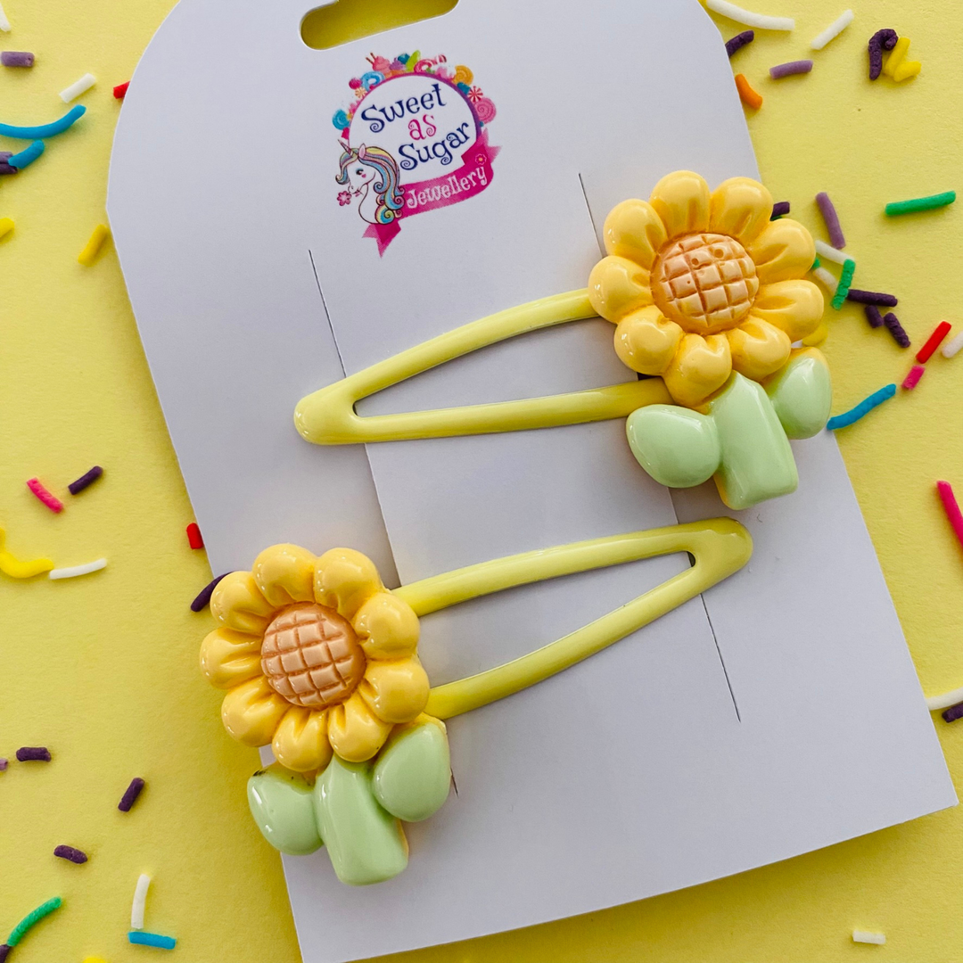 A pair of yellow snap clips featuring a yellow sunflower with green stem charm.